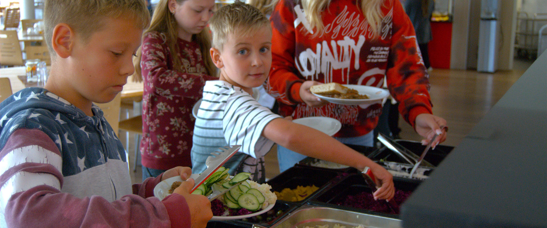 Kinder servieren sich an einem Buffet; hell-dunkel gestreifte Kleidung, konzentrierte Ausdrücke.