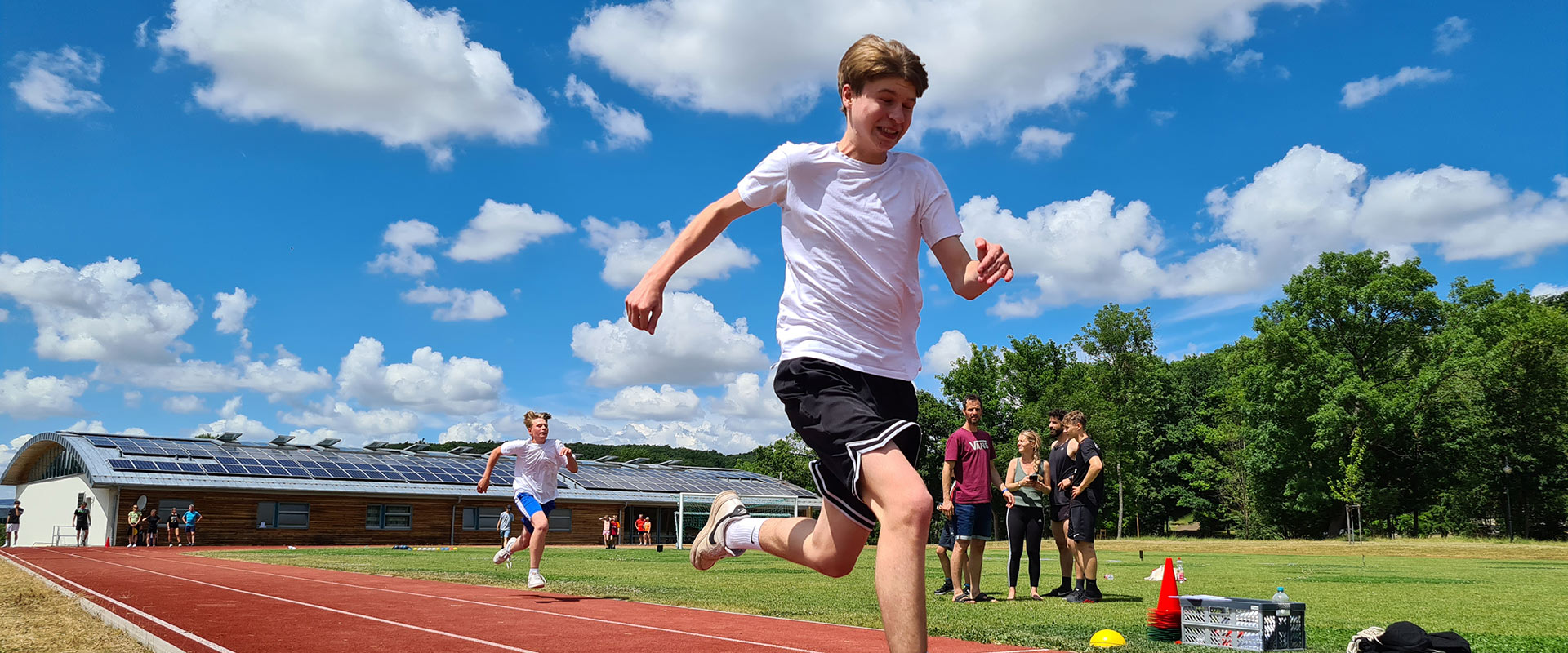 Junger Mann lacht, läuft auf roter Bahn; blau-weißer Himmel, grüner Hintergrund, Zuschauer dahinter.