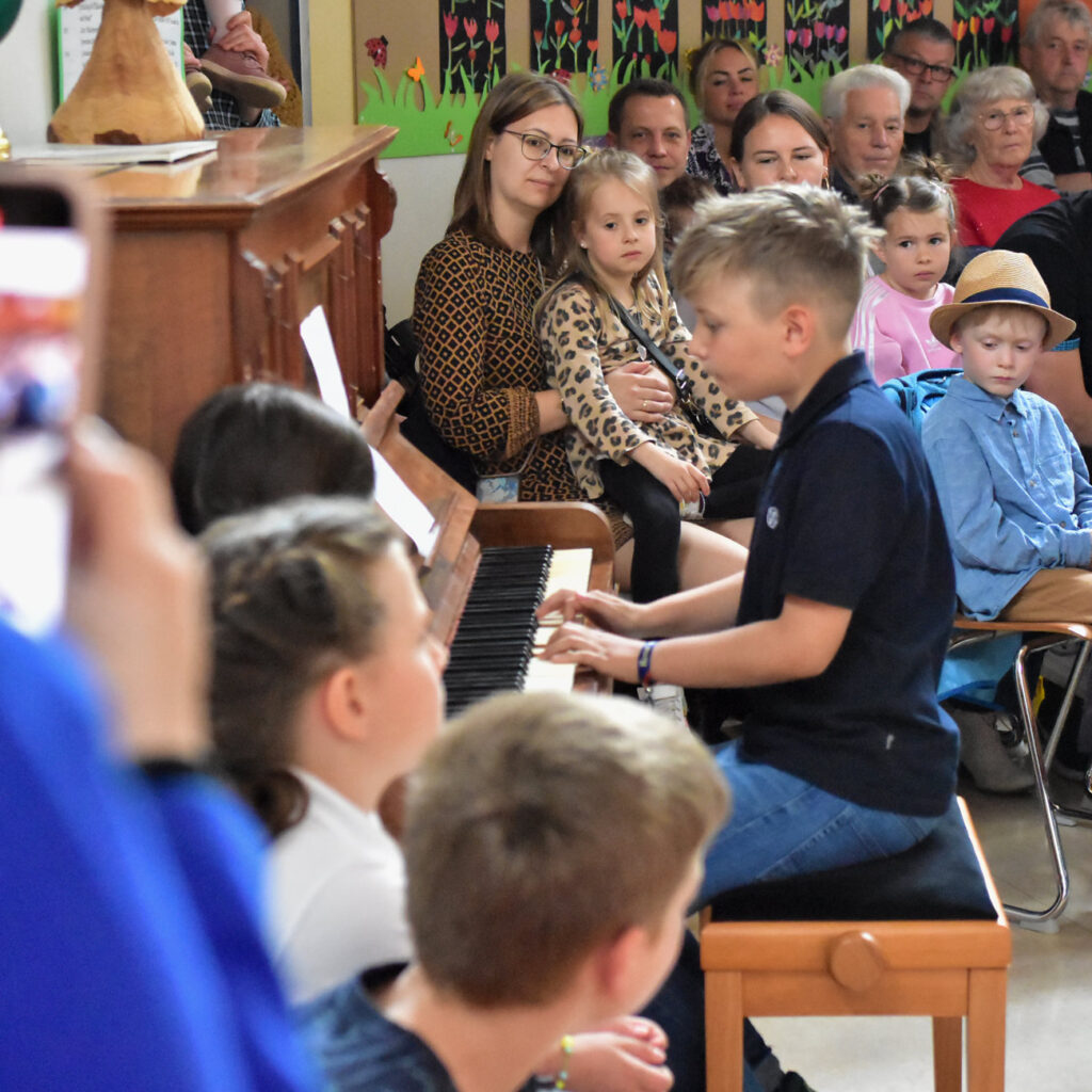 A boy is playing the piano, surrounded by adults and children, in a room with colorful walls.