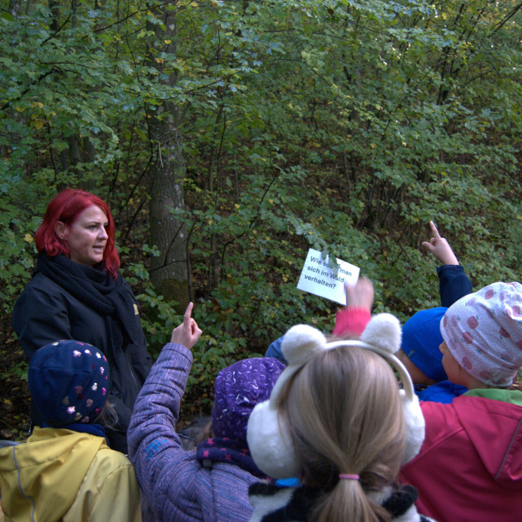 Eine Person mit rotem Haar spricht lächelnd zu einer Gruppe von Kindern im bunten Herbstwald.