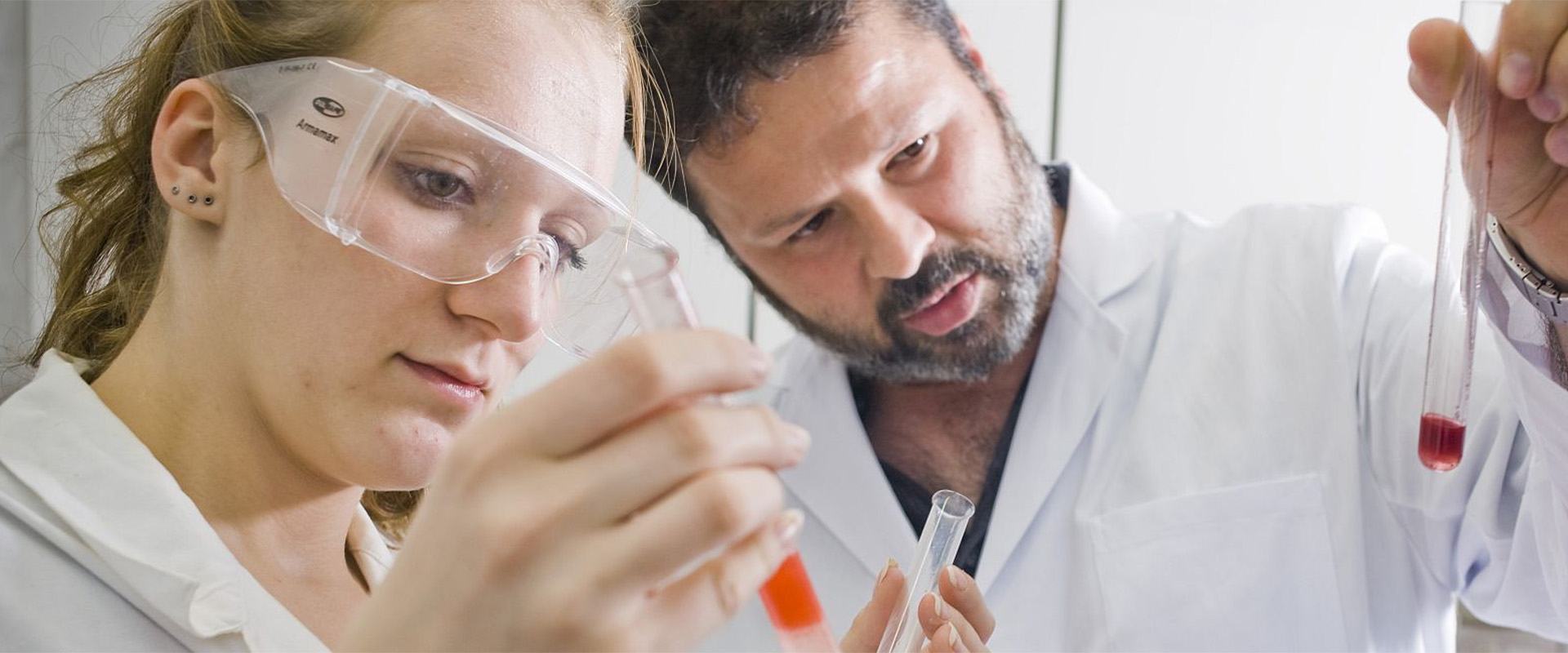 Two people in lab coats, looking focused, with test tubes and red liquid.