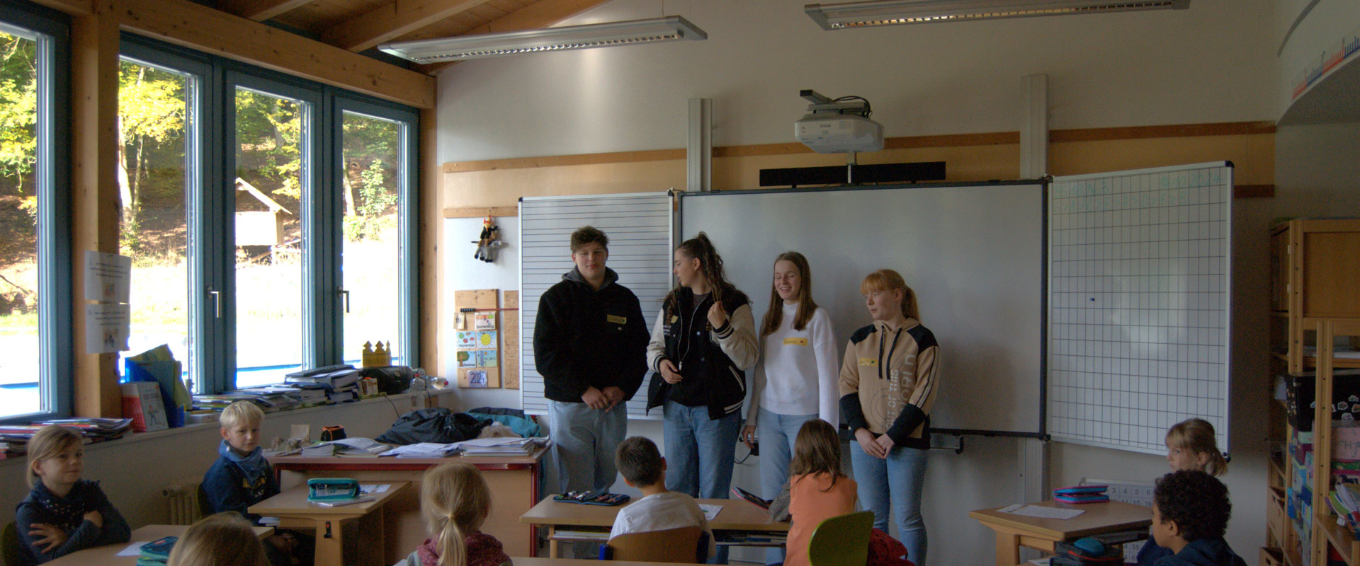 Vier lächelnde junge Erwachsene präsentieren vor Grundschulkindern in einem hellen Klassenraum.