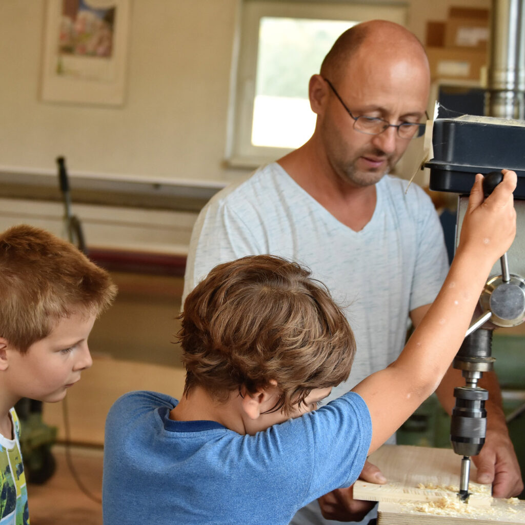 A man is supervising two children as they drill. Everyone is concentrating. The background is out of focus.