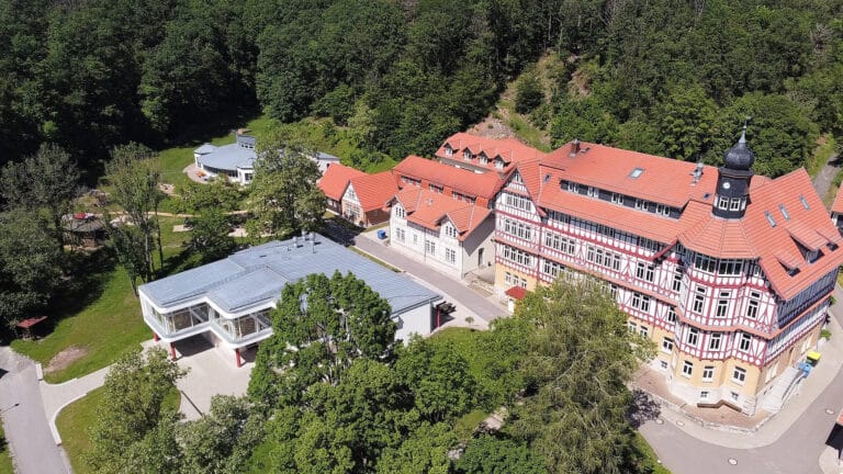 Luftaufnahme eines großen Gebäudes mit roten Dächern, umgeben von Wald, bei sonnigem Wetter.