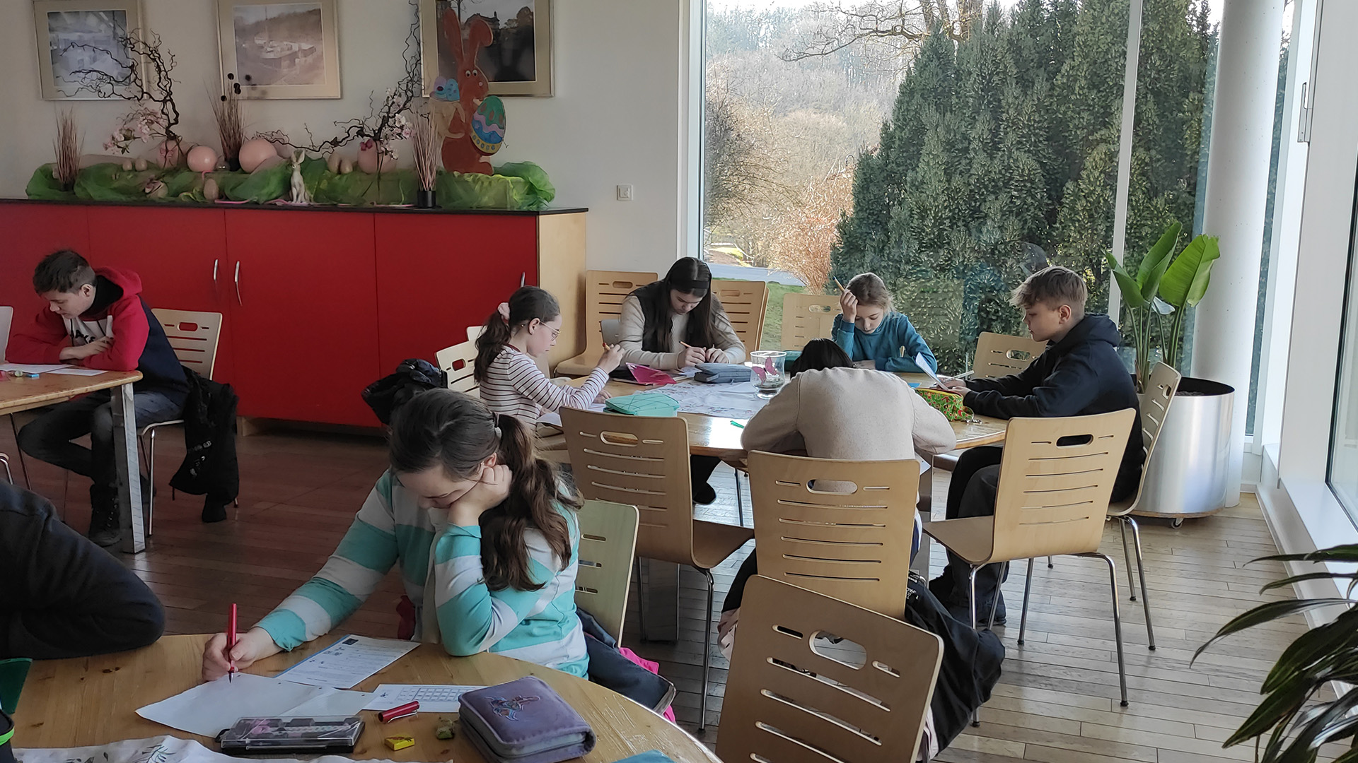 Mehrere Kinder konzentrieren sich beim Schreiben in einem sonnendurchfluteten Raum mit roten Möbeln.