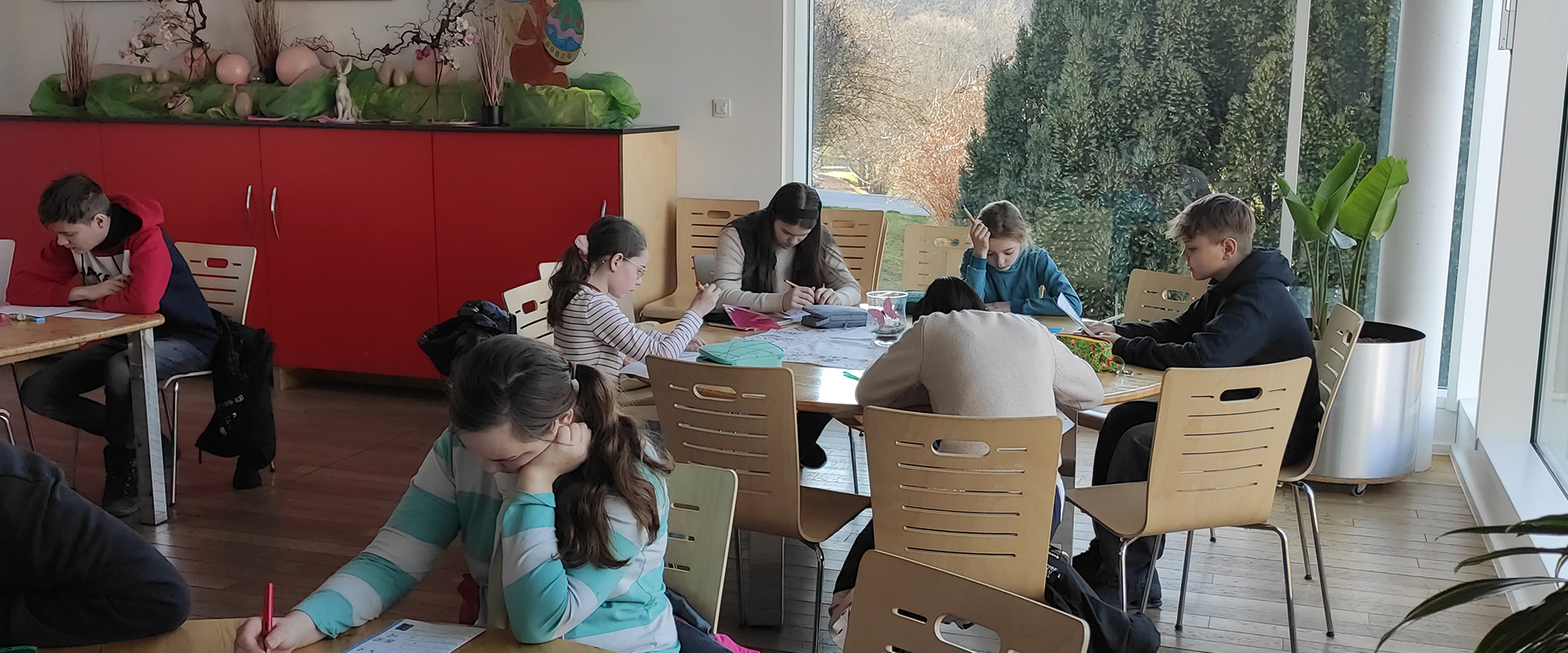 Schüler:innen an Tischen, konzentriert schreibend, helle Fensteransicht, rote Schränke im Hintergrund.