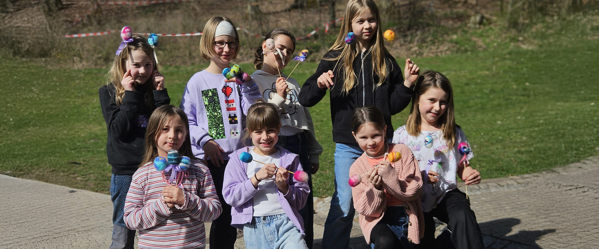 Eight smiling children holding colorful pom-poms in front of a grassy landscape.