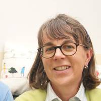 Eine lächelnde Frau mit Brille und braunem Haar trägt einen grünen Pullover.