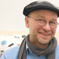 Ein lächelnder Mann mit Brille, schwarzer Mütze und Schal vor hellem Hintergrund