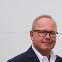 Ein Mann mit Brille lächelt leicht, trägt einen dunklen Anzug und weißes Hemd
