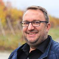 Ein lächelnder Mann mit Brille in blauer Jacke vor herbstlicher Kulisse