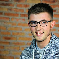 Ein lächelnder Mann mit Brille vor einer Ziegelwand, trägt einen Hoodie