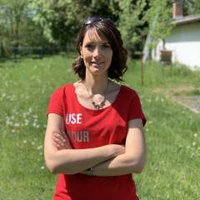 Frau, lächelnd, braunes Haar, rotes Shirt, steht draußen im Sonnenschein