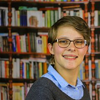Eine lächelnde, brünette Person mit Brille steht vor einem Bücherregal.