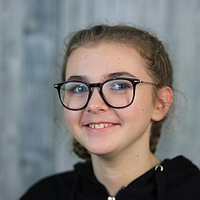 Eine junge Person mit Brille lächelt freundlich, dunkler Hoodie, grauer Hintergrund