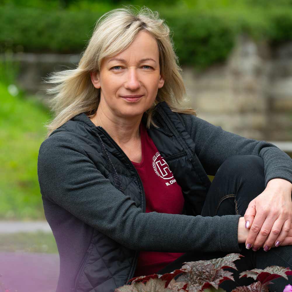 A woman with blonde hair is sitting outside, smiling, and wearing a black jacket.