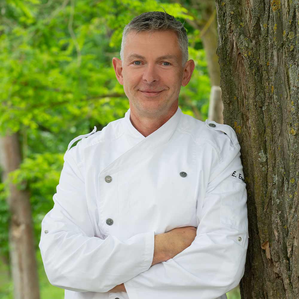 A smiling man with gray hair, wearing a white chef's coat, is leaning against a tree.