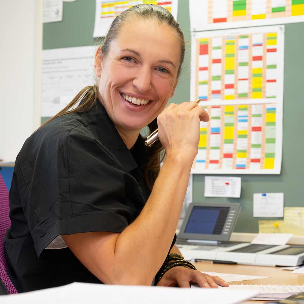 A smiling woman in a dark shirt is sitting at a desk covered with papers.