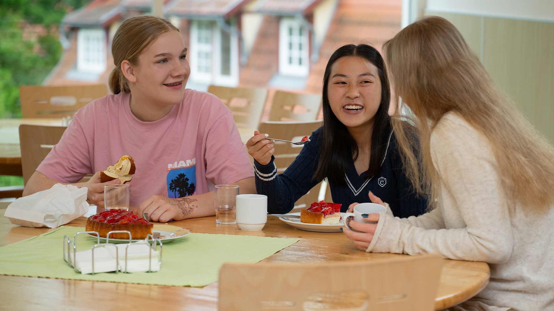 Drei junge Frauen lachen gemeinsam am Tisch und essen Kuchenstücke.