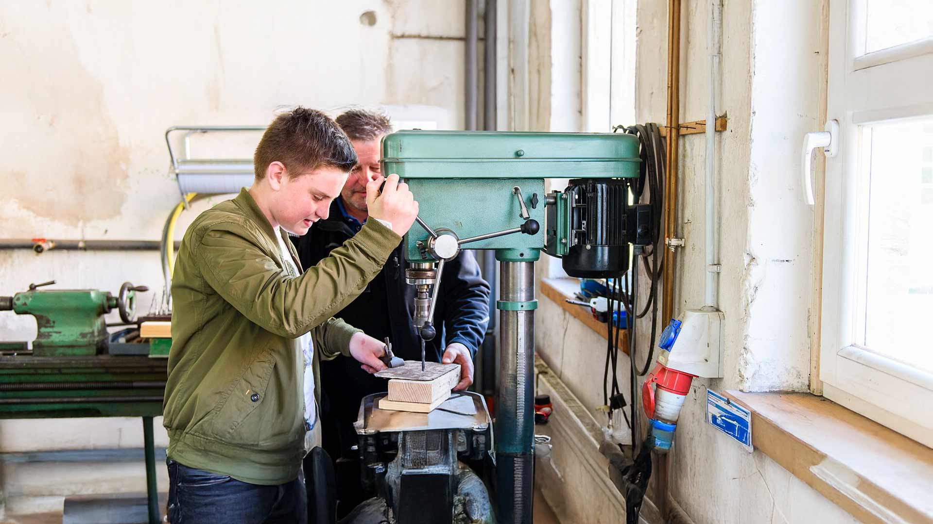Zwei Personen verwenden eine grüne Bohrmaschine in einer Werkstatt mit hellem Licht.