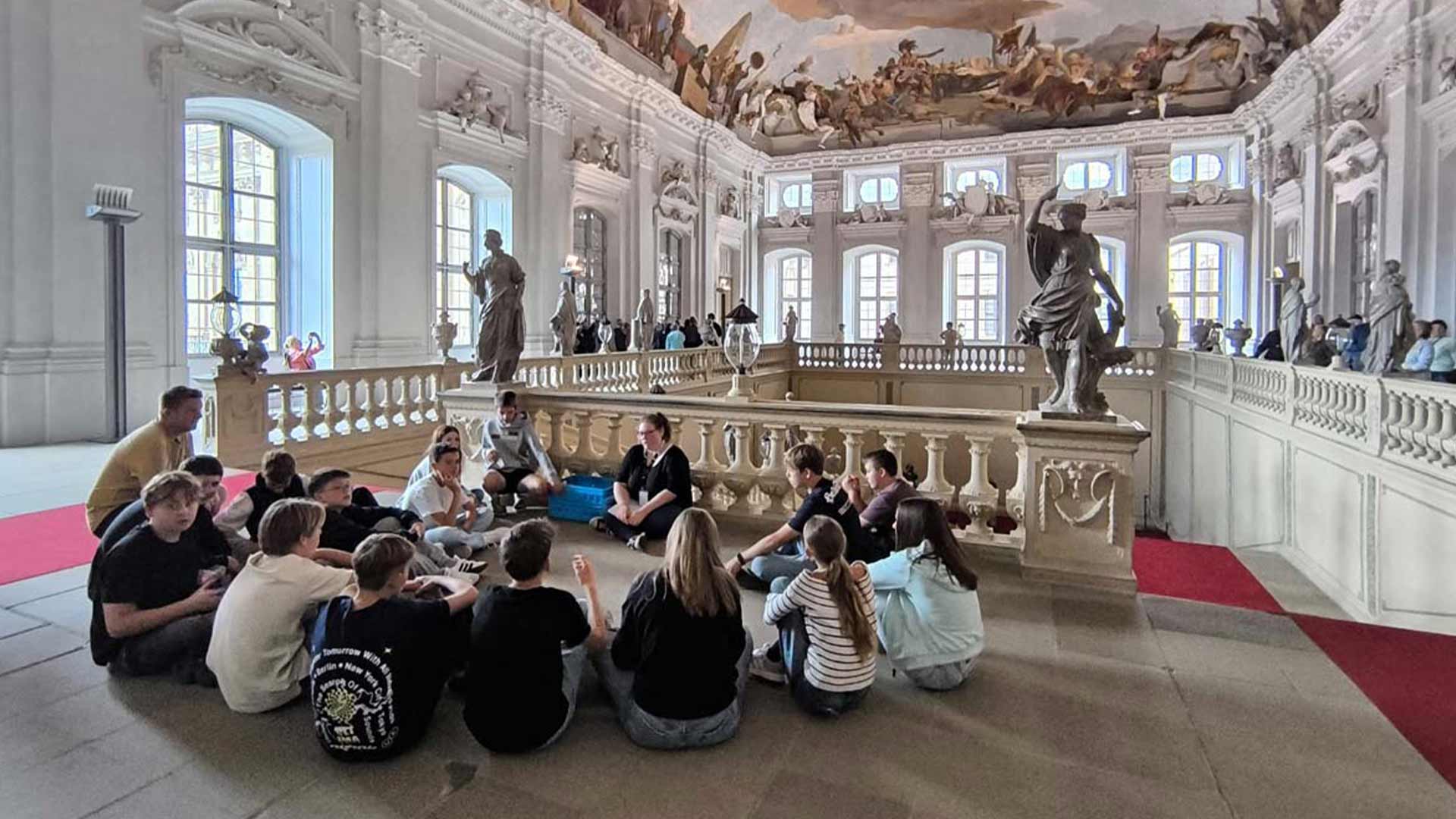 Eine Gruppe von Menschen sitzt auf dem Boden in einem prächtigen, kunstvoll dekorierten Raum mit Statuen und Wandmalereien.