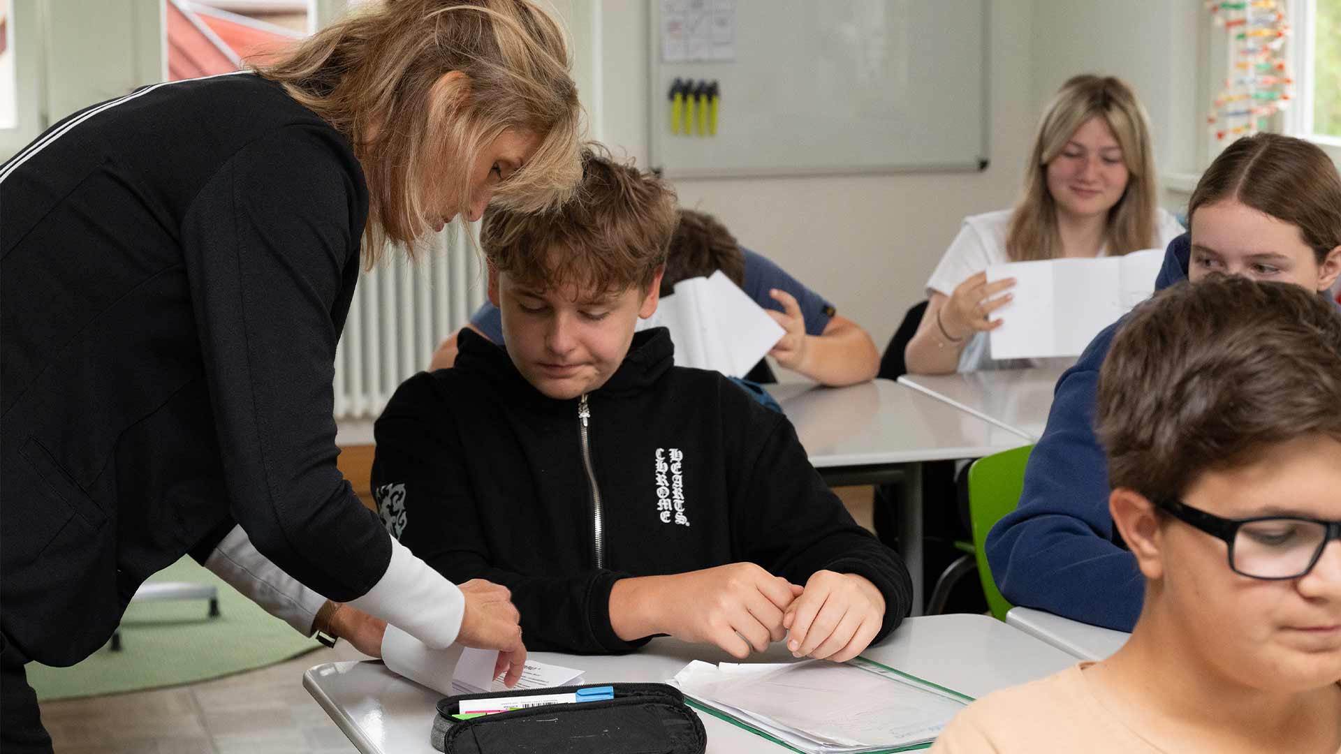 Eine Lehrerin hilft einem Schüler in einem belebten Klassenzimmer mit Aufgaben.
