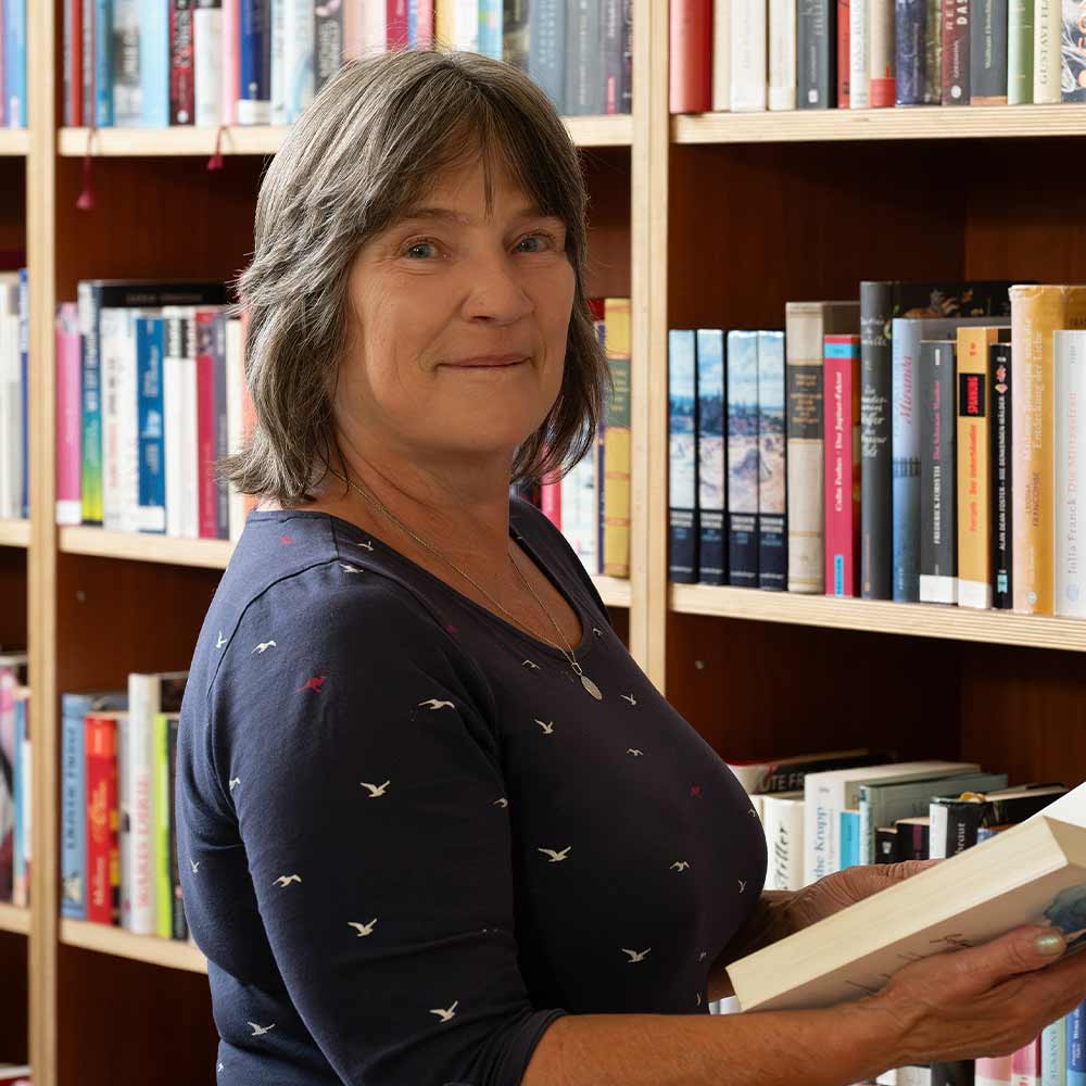 A woman with brown hair is standing in a library, smiling and holding a book.