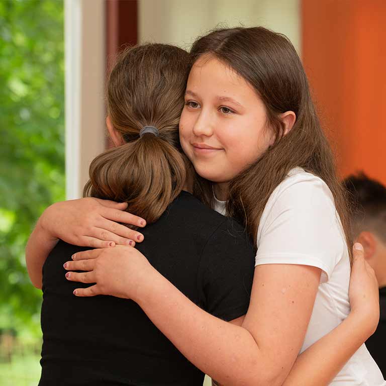 A young girl with brown hair is hugging another girl; both are smiling.