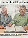 Two older men in an office; one is smiling, the other looks serious; there are documents on the table.