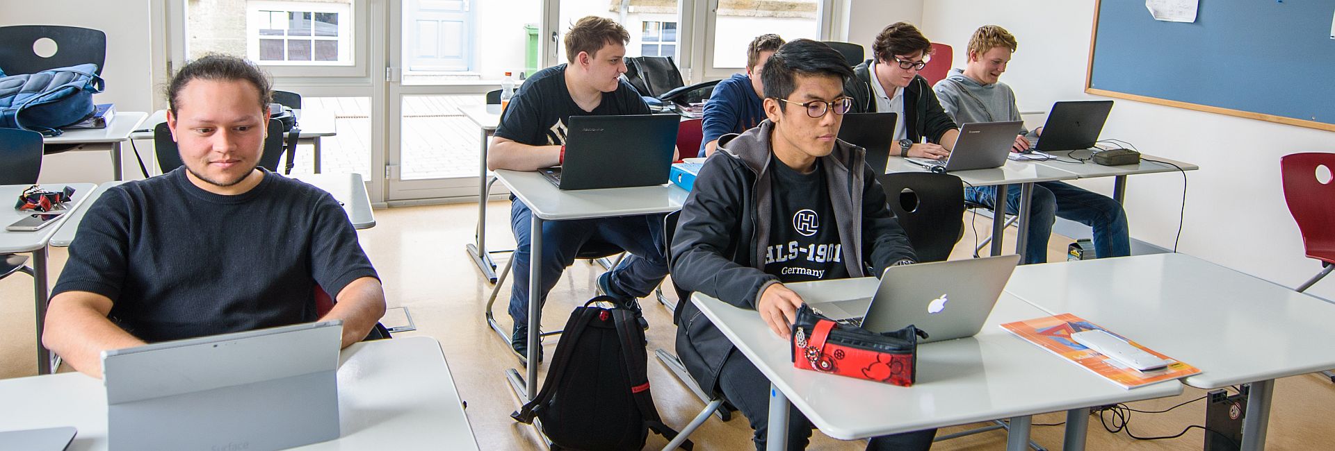 Mehrere junge Menschen sitzen konzentriert an Laptops in einem Klassenzimmer.