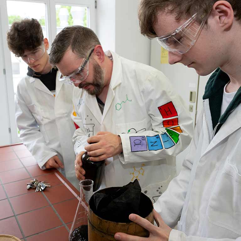 Drei Personen in weißen Laborkitteln arbeiten konzentriert mit Glasflaschen und Messbechern.