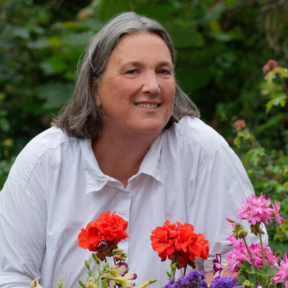 A smiling person with gray hair wearing a white shirt, standing in front of colorful flowers.