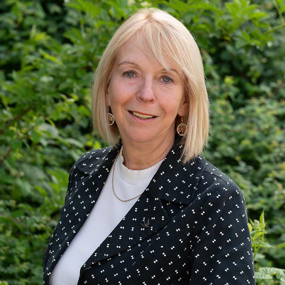 A smiling woman with blonde hair is wearing a dark patterned blazer.