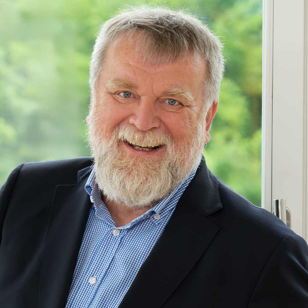A smiling older man with a beard, wearing a blue shirt and a dark jacket, is standing in front of a window overlooking a green landscape.