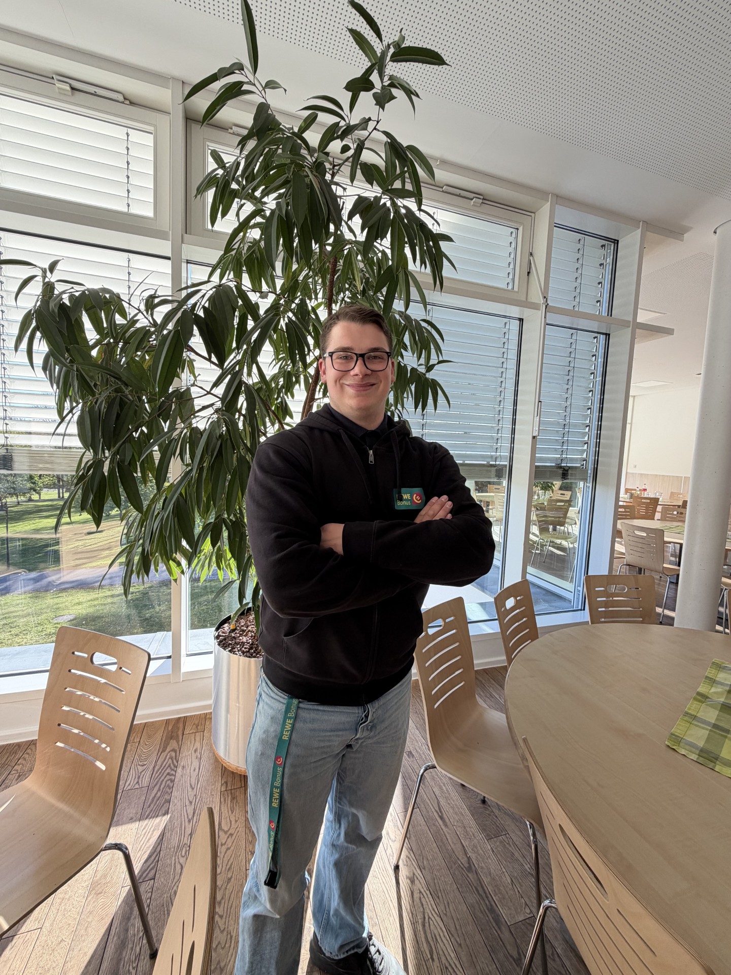 Ein junger Mann mit Brille lächelt entspannt in heller Cafeteria neben grünem Baum