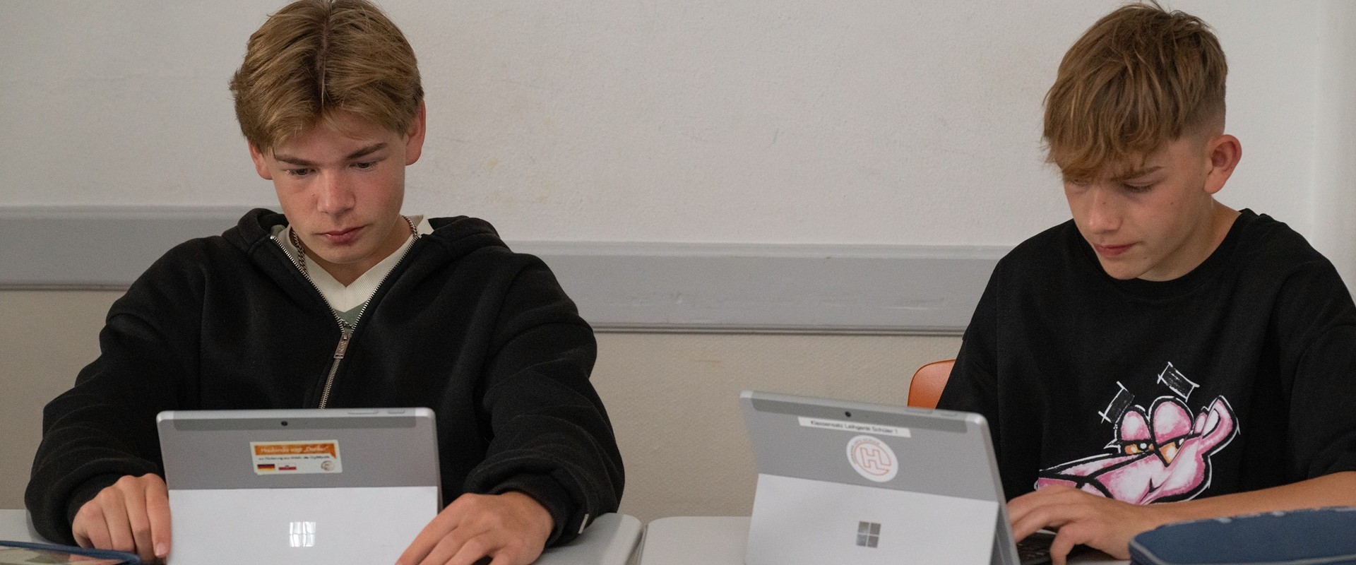 Two teenagers with serious expressions are working intently on silver tablets in the classroom