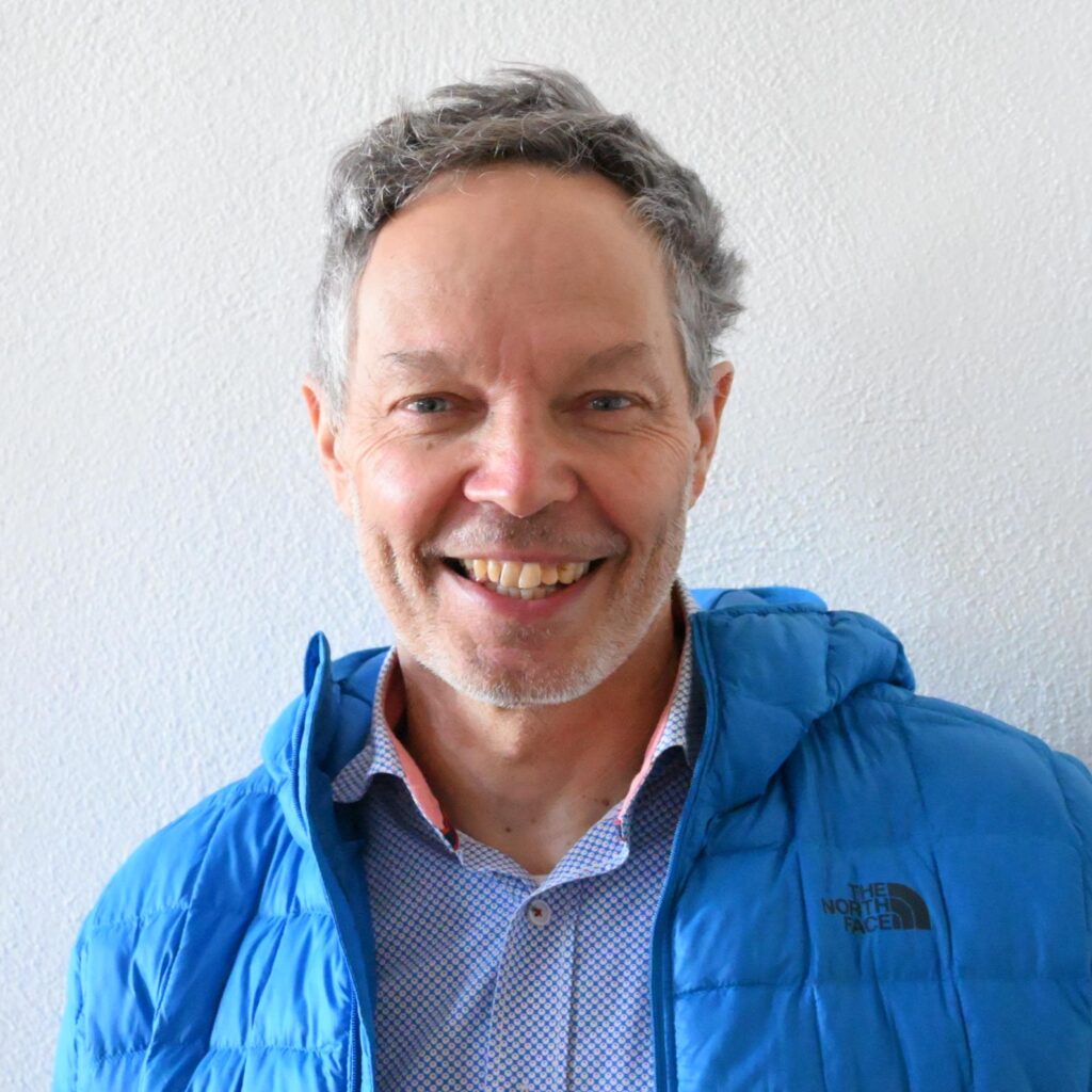 A smiling older man with gray hair wearing a blue jacket in front of a white wall