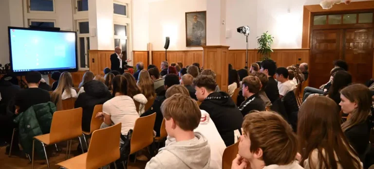 Mehrere Schüler hören aufmerksam einem älteren Mann vor Bildschirm zu