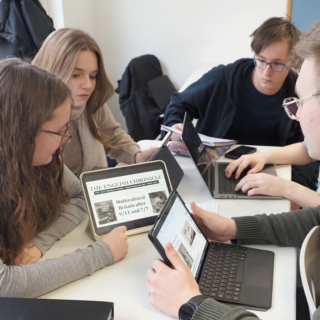 Four young people with neutral expressions are working on laptops at a table