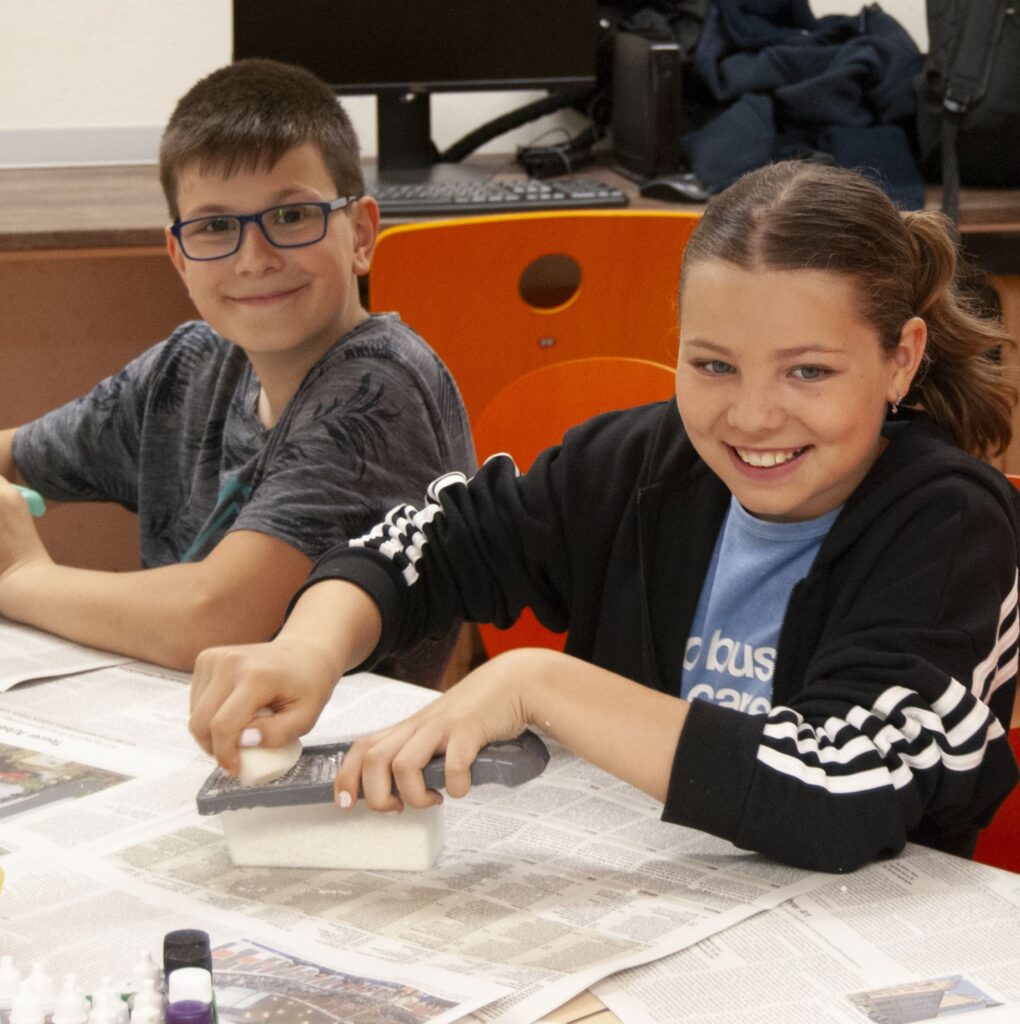 Two children wearing glasses and pigtails smile cheerfully as they do crafts at the table