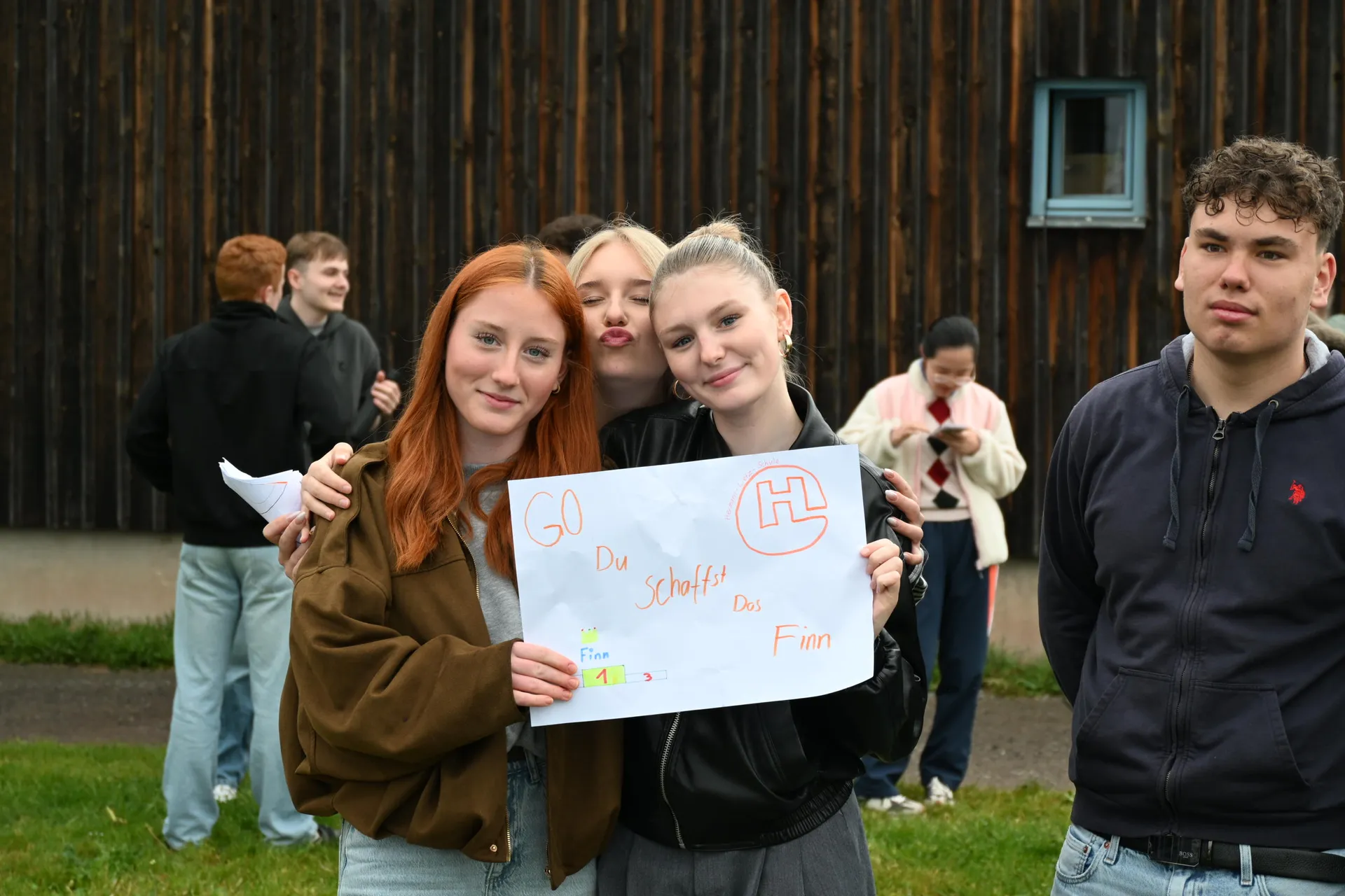 Drei junge Menschen lächeln mit einem handgemalten Schild vor einer Holzwand