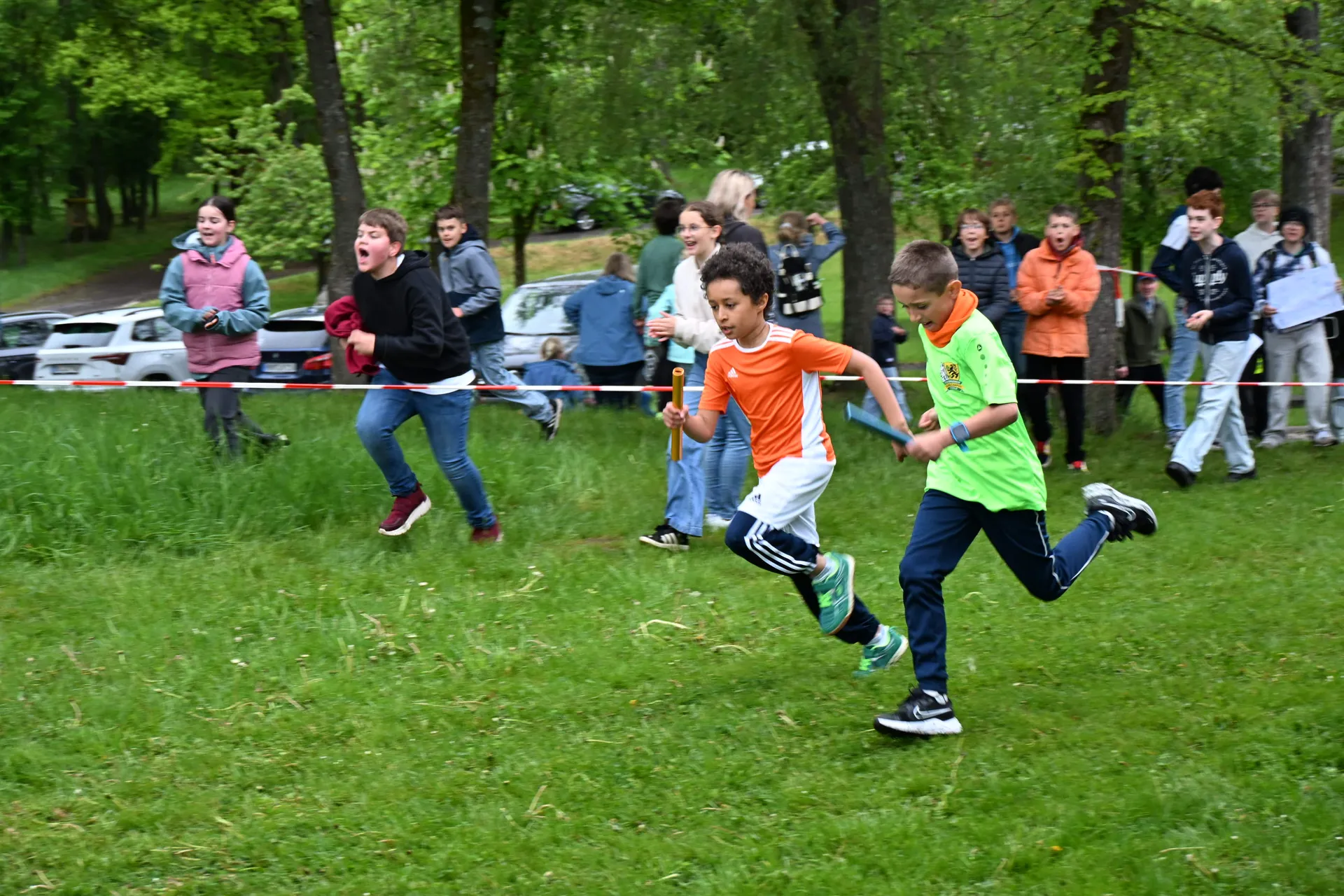 Drei Jungen laufen lachend im Freien, zwei tragen orange und grüne Sportkleidung