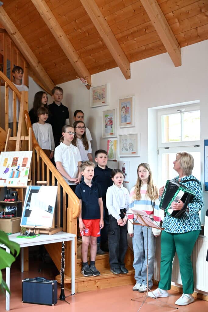 Eine Gruppe von Kindern singt mit freundlicher Miene auf einer Holztreppe neben einer Frau in grün weißer Kleidung, die ein Akkordeon spielt