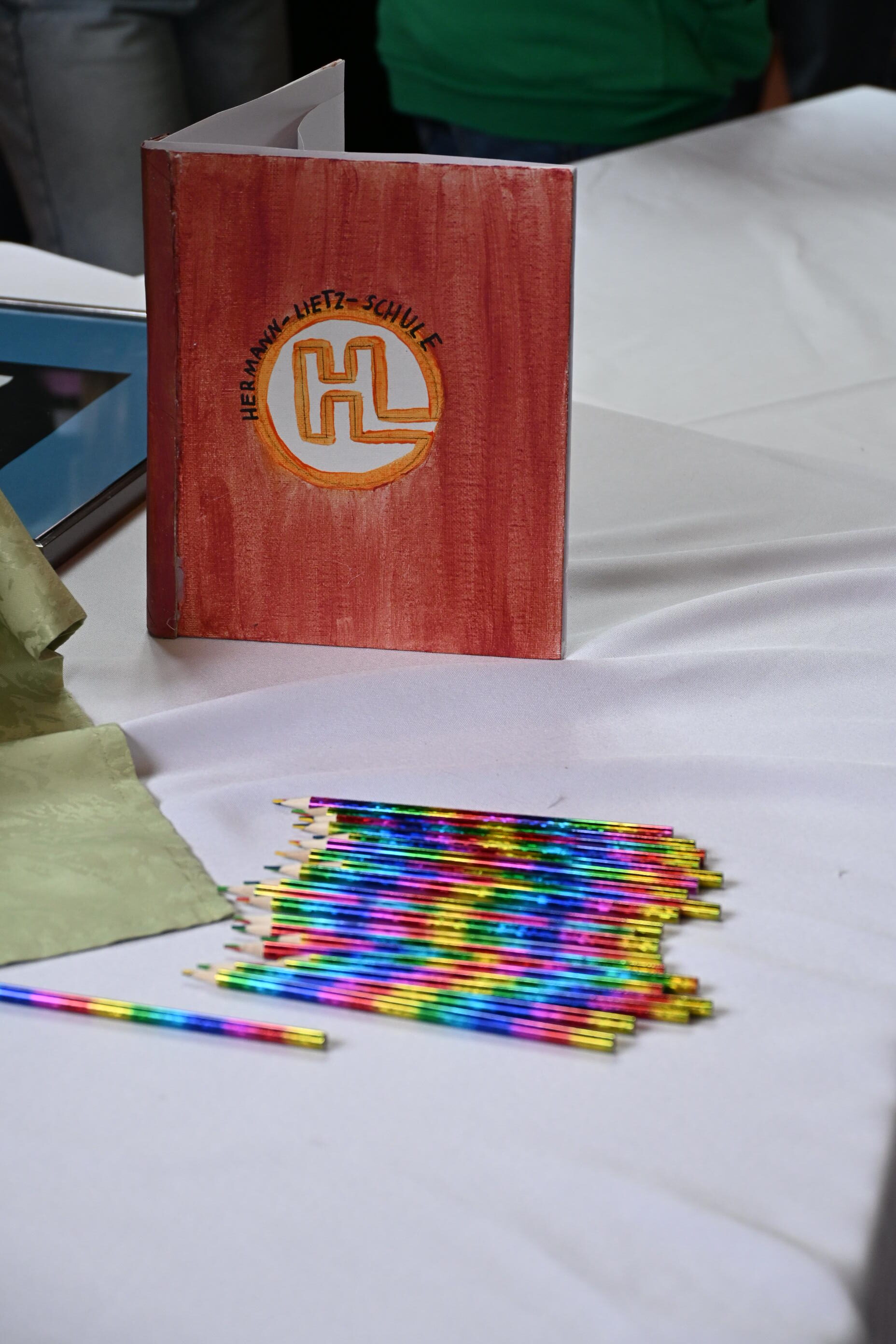Ein aufgeklapptes rotes Buch mit Logo sowie bunte Stifte auf weißem Tisch