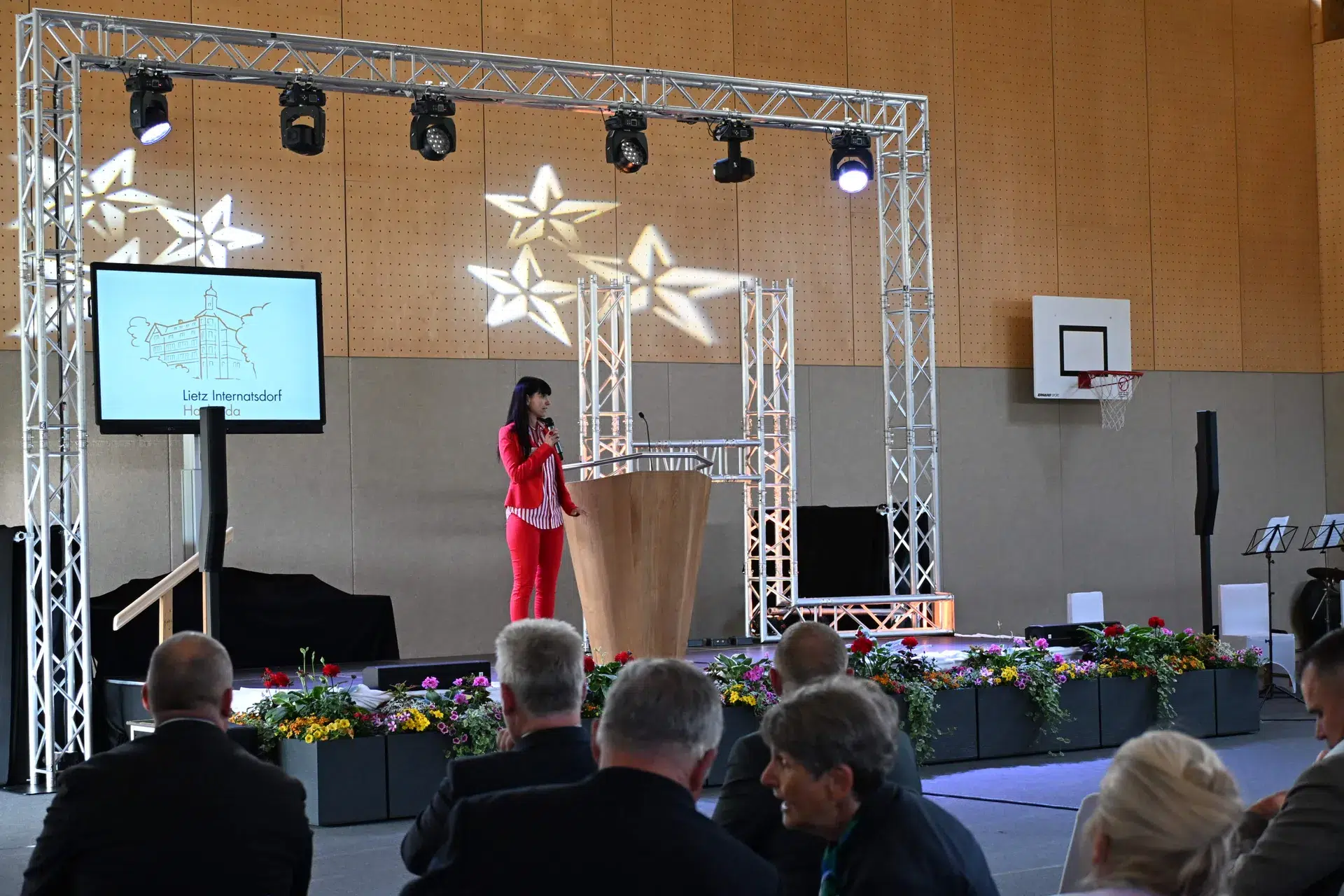 Eine Frau in rotem Anzug spricht lächelnd am Podium vor Publikum in Halle