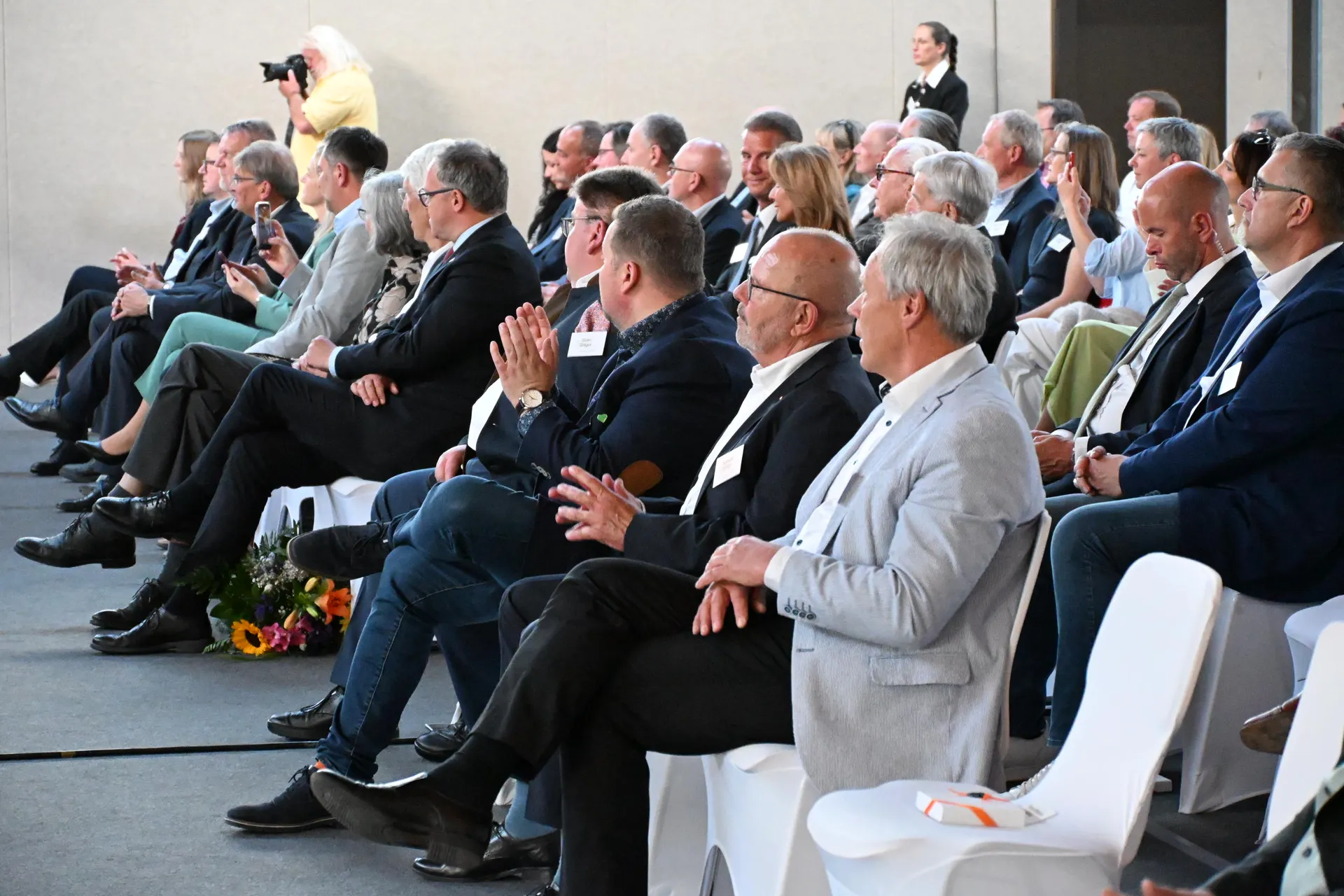 Mehrere Personen in feiner Kleidung sitzen aufmerksam auf weißen Stühlen im Saal