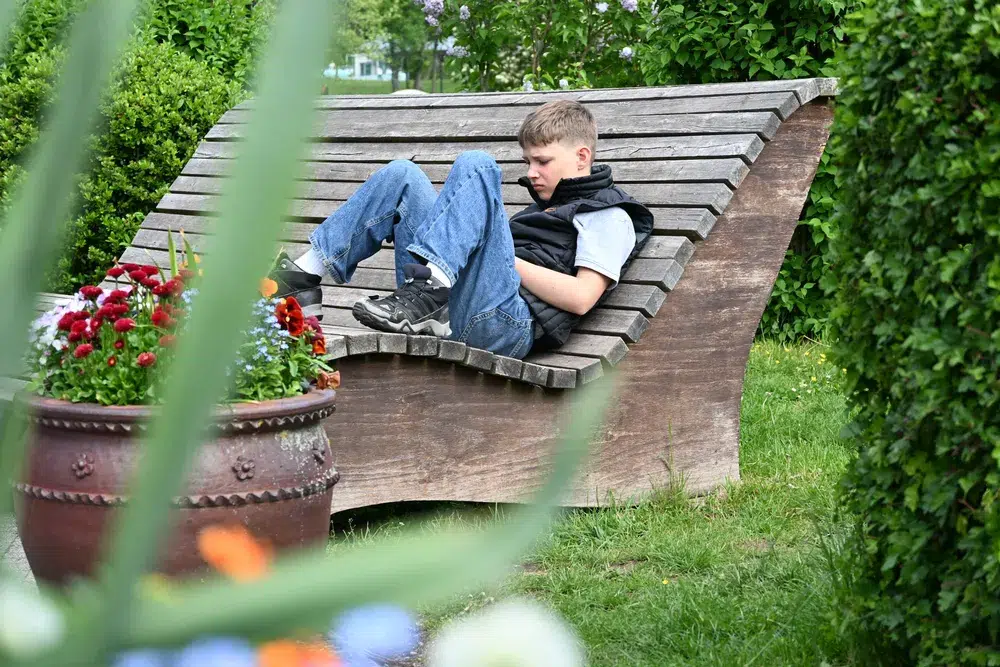 Junge mit entspanntem Gesichtsausdruck liegt auf einer Holzbank im Grünen
