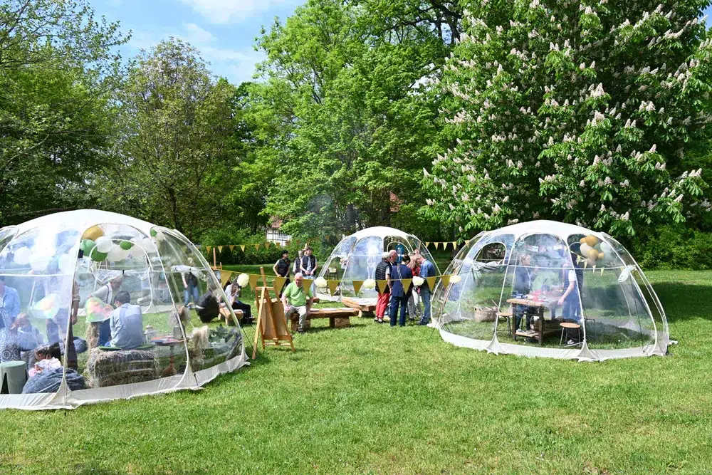Mehrere Personen stehen lächelnd im Grünen um transparente Kuppeln mit bunten Dekorationen