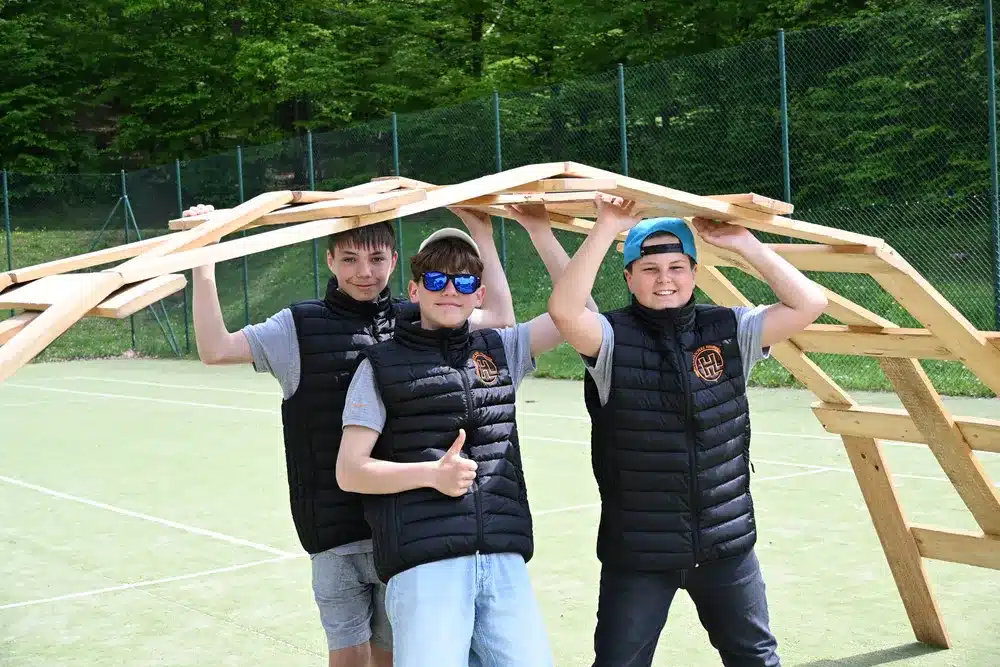 Drei junge Männer stehen lachend unter einer Holzkonstruktion auf grünem Sportplatz
