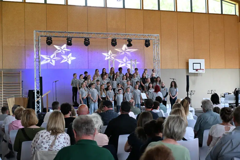 Eine Gruppe von Kindern und Jugendlichen singt fröhlich vor einem Publikum in einer hellen Turnhalle